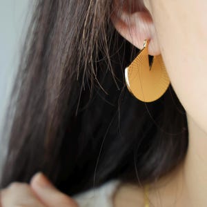 May include: Close-up of a gold-colored earring with a fan-like design. The earring is a half-circle shape with a cut-out section at the top. The surface of the earring has a textured, radial pattern. The earring is worn on a person's ear.