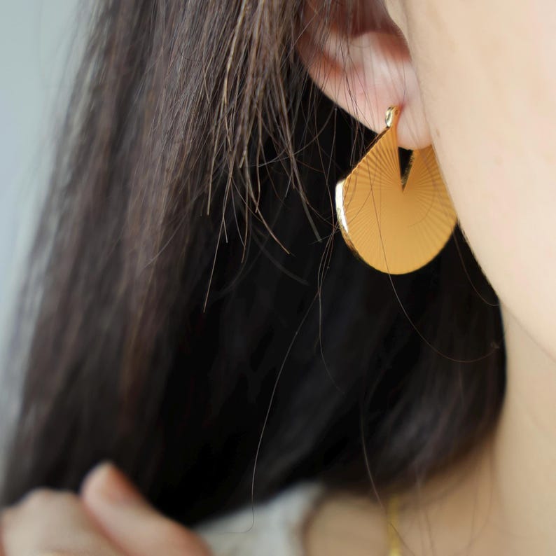 May include: Close-up of a gold-colored earring with a fan-like design. The earring is a half-circle shape with a cut-out section at the top. The surface of the earring has a textured, radial pattern. The earring is worn on a person's ear.