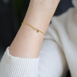 May include: A delicate gold chain bracelet with a small, round charm. The bracelet is shown on a person's wrist.