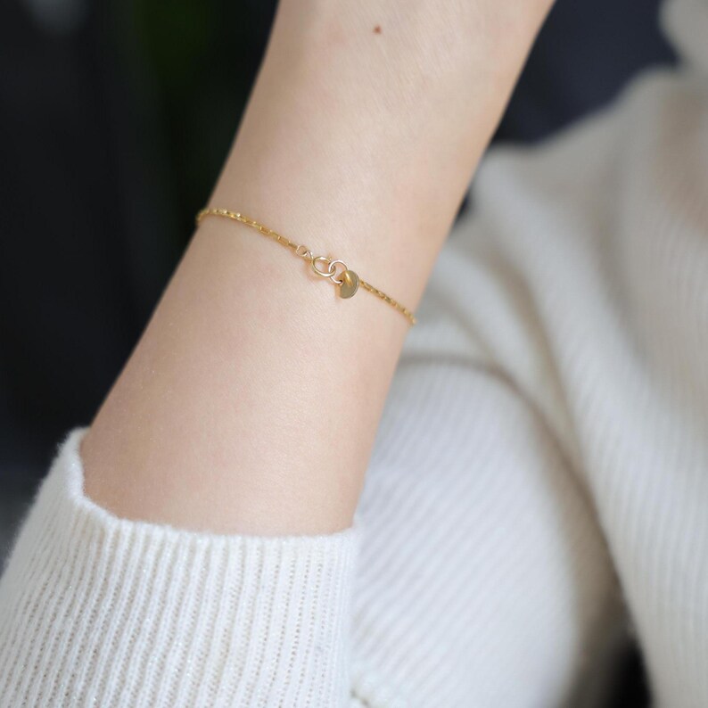 May include: A delicate gold chain bracelet with a small, round charm. The bracelet is shown on a person's wrist.
