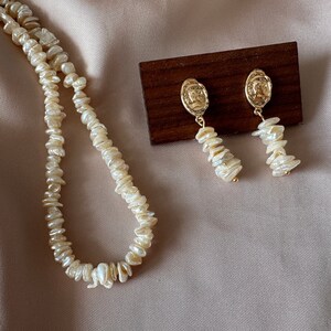 Puede incluir: Un conjunto de collar y pendientes de color crema. El collar est&aacute; hecho de perlas de forma irregular. Los pendientes tienen un medall&oacute;n dorado con un perfil y una hebra de perlas. Las joyas se exhiben sobre un fondo neutro.