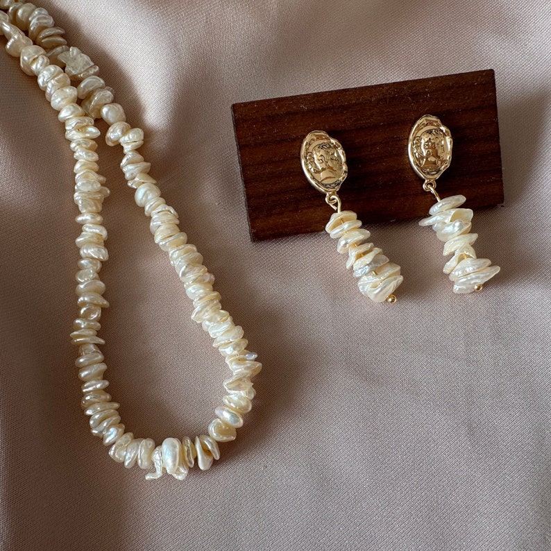 Puede incluir: Un conjunto de collar y pendientes de color crema. El collar est&aacute; hecho de perlas de forma irregular. Los pendientes tienen un medall&oacute;n dorado con un perfil y una hebra de perlas. Las joyas se exhiben sobre un fondo neutro.