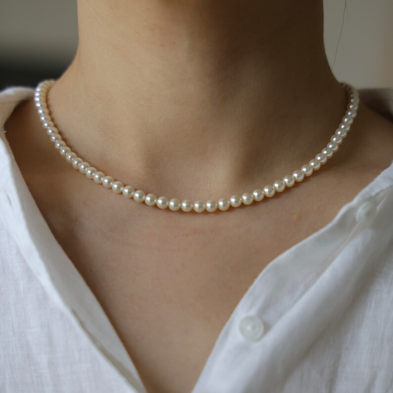 May include: A delicate pearl necklace composed of small, round, off-white pearls. The necklace is worn against a white shirt, creating a classic and elegant look. The pearls are evenly spaced, forming a continuous strand.