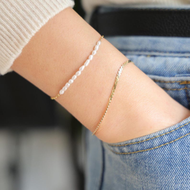 May include: Two bracelets on a person's wrist. The top bracelet is a delicate chain with small white pearls. The bottom bracelet is a gold chain with a slightly textured finish.