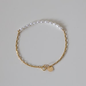 May include: A delicate gold chain bracelet with a row of white pearls on the top half. The bracelet has a heart-shaped clasp.
