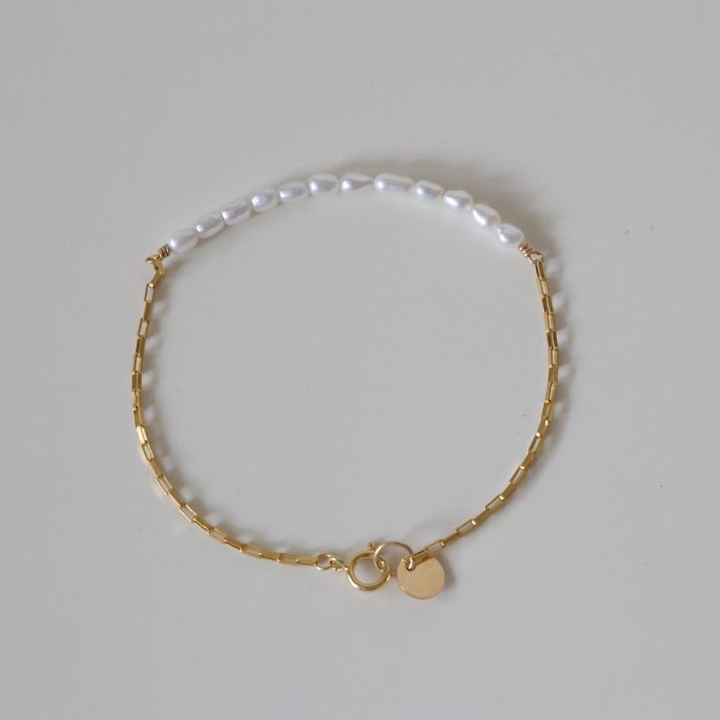 May include: A delicate gold chain bracelet with a row of white pearls on the top half. The bracelet has a heart-shaped clasp.