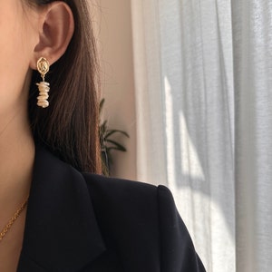 Puede incluir: Pendientes colgantes dorados con un peque&ntilde;o dise&ntilde;o circular con un emblema detallado. Los pendientes tienen un dise&ntilde;o de perlas irregulares colgantes. La imagen muestra los pendientes con una blazer negra y un collar dorado.