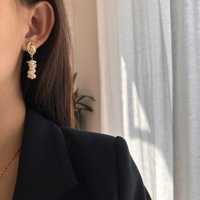 Puede incluir: Pendientes colgantes dorados con un peque&ntilde;o dise&ntilde;o circular con un emblema detallado. Los pendientes tienen un dise&ntilde;o de perlas irregulares colgantes. La imagen muestra los pendientes con una blazer negra y un collar dorado.