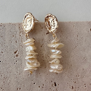 Puede incluir: Pendientes colgantes dorados con un dise&ntilde;o de moneda en la parte superior y una cascada de perlas irregulares de color blanco roto. Los pendientes tienen cierre de poste y una peque&ntilde;a cuenta dorada en la parte inferior.