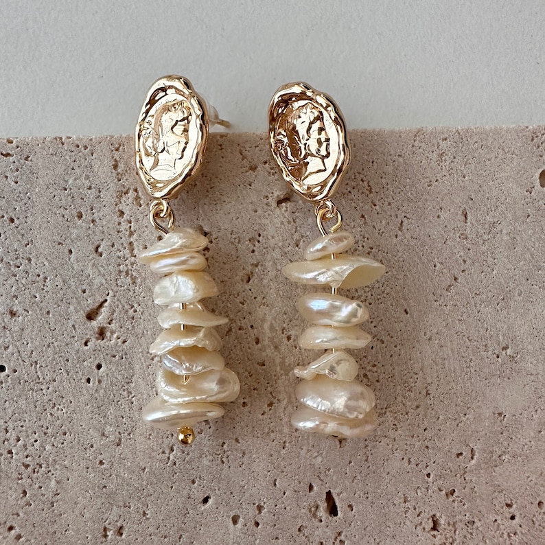 Puede incluir: Pendientes colgantes dorados con un dise&ntilde;o de moneda en la parte superior y una cascada de perlas irregulares de color blanco roto. Los pendientes tienen cierre de poste y una peque&ntilde;a cuenta dorada en la parte inferior.