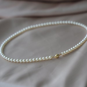 May include: A delicate pearl necklace with a gold clasp. The necklace is composed of small, round, white pearls and is displayed on a soft, neutral-toned fabric. The gold clasp adds a touch of elegance.