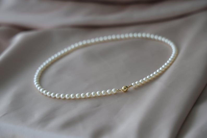May include: A delicate pearl necklace with a gold clasp. The necklace is composed of small, round, white pearls and is displayed on a soft, neutral-toned fabric. The gold clasp adds a touch of elegance.