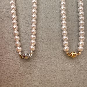 May include: Two strands of cream-colored pearl necklaces. One necklace has a silver clasp, and the other has a gold clasp. The pearls are round and evenly spaced. The background is a neutral gray.
