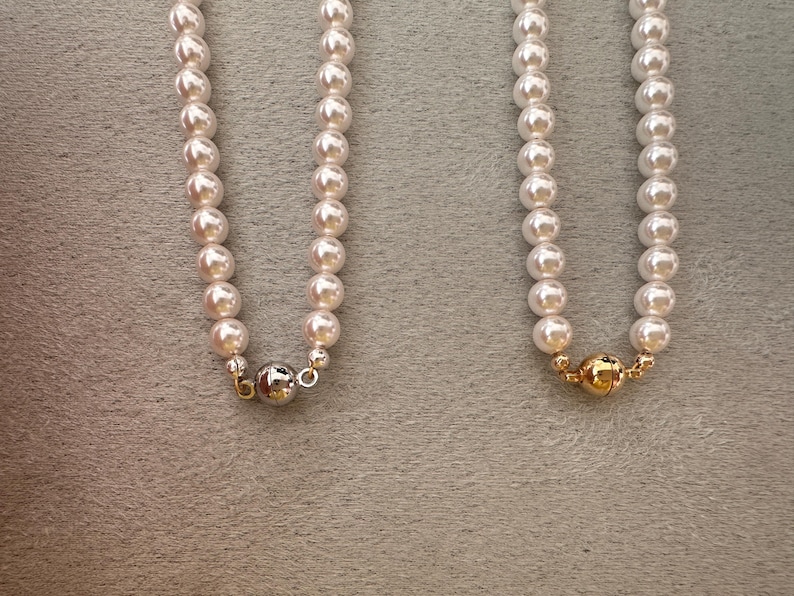 May include: Two strands of cream-colored pearl necklaces. One necklace has a silver clasp, and the other has a gold clasp. The pearls are round and evenly spaced. The background is a neutral gray.