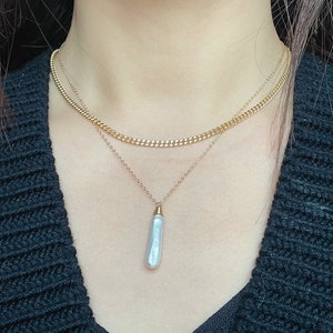 May include: A layered gold chain necklace with a single, long, white pearl pendant.