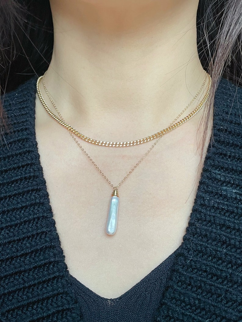 May include: A layered gold chain necklace with a single, long, white pearl pendant.