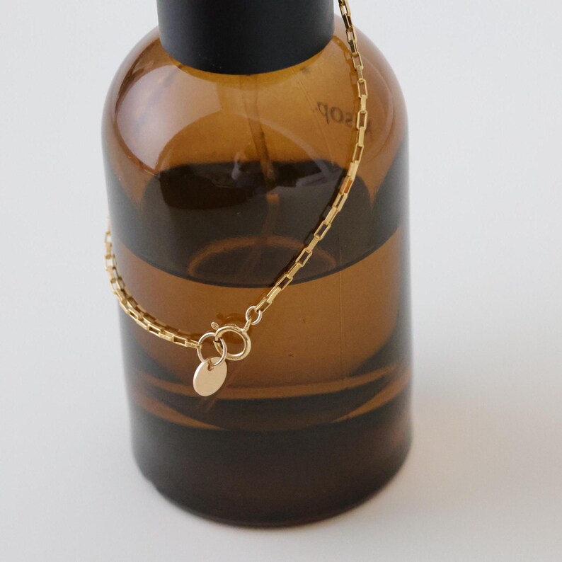 May include: A gold chain bracelet with a small round charm. The bracelet is hanging from a brown glass bottle.