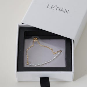 May include: A delicate gold chain bracelet with a row of small white pearls. The bracelet is displayed in a white box with the word "LE'TIAN" printed on the lid.