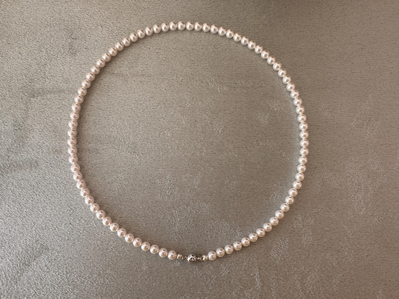 May include: A delicate pearl necklace with a silver clasp. The necklace is composed of numerous small, round, white pearls, strung together to form a complete circle. The clasp is a small, silver-colored metal piece. The necklace is displayed on a gray surface.