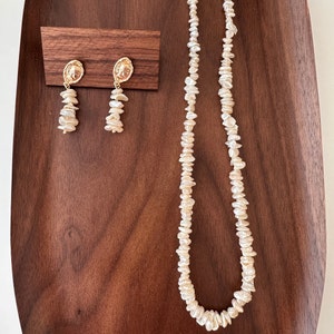 Puede incluir: Un conjunto de joyas con collar y pendientes. El collar y los pendientes est&aacute;n hechos de peque&ntilde;as cuentas irregulares de color blanco roto. Los pendientes tienen tapas ovaladas doradas con un dise&ntilde;o en relieve. Las joyas se exhiben sobre una superficie de madera oscura.