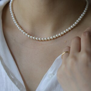 May include: A delicate pearl necklace with small, round, off-white beads. The necklace is worn against a white collared shirt. A gold ring is visible on a finger.