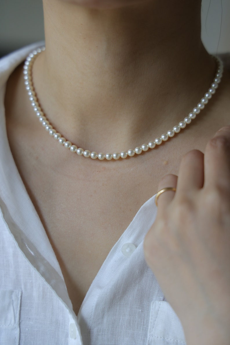 May include: A delicate pearl necklace with small, round, off-white beads. The necklace is worn against a white collared shirt. A gold ring is visible on a finger.