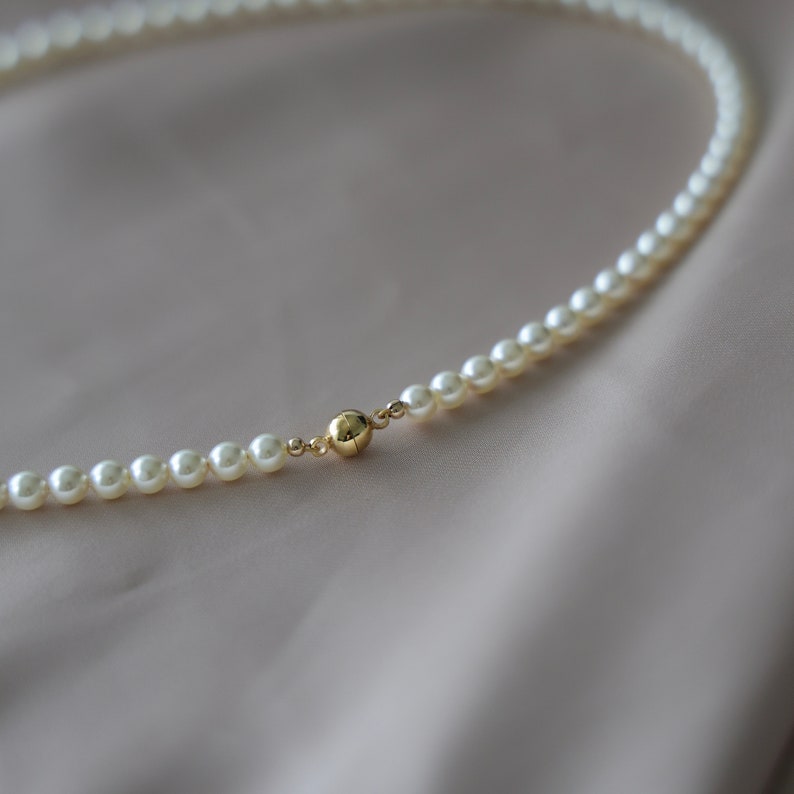 May include: A delicate pearl necklace with a gold clasp. The necklace features small, round, off-white pearls strung together. The gold clasp is a small, round sphere. The necklace is displayed on a soft, neutral-toned fabric.