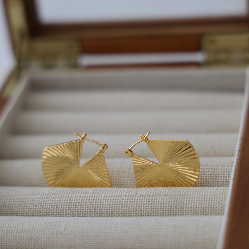May include: A pair of gold-colored, geometric hoop earrings. Each earring has a square shape with a fan-like textured design. The earrings are displayed inside a wooden jewelry box with a cream-colored interior.