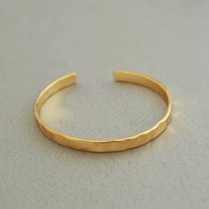 Könnte beinhalten: Ein goldfarbenes offenes Armband mit strukturierter Oberfläche. Das Armband besteht aus glänzendem, gelbem Metall und hat ein schlichtes, minimalistisches Design. Das Armband ist an den Enden offen und zum Tragen am Handgelenk bestimmt.