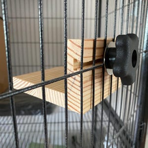 May include: A wooden shelf with a black knob attached to a metal cage. The shelf is made of light wood and has a simple design.