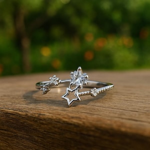 Puede incluir: Un anillo plateado con un diseño de estrella y floral. El anillo presenta una estrella abierta y está adornado con pequeñas piedras preciosas brillantes. El anillo está sobre una superficie de madera con un fondo verde borroso.