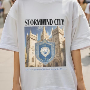May include: White t-shirt with a graphic print of a castle and a lion crest. The text "Stormwind City" is printed above the graphic. The text below the graphic reads "A bastion of hope and the last hope of the Alliance."