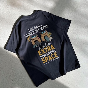 May include: Black t-shirt with a graphic of two cartoon bags with eyes and the text "The bags under my eyes are for extra inventory space".