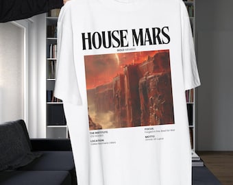 May include: White t-shirt with a graphic print of a red and orange landscape with the text "HOUSE MARS" in black. The text "GOLD MEMBER" is printed below the graphic. The text "THE INSTITUTE 1/2 HOUSES" is printed below the graphic. The text "LOCATION Valles Marineris | Mars" is printed below the graphic. The text "FOCUS Forged in Fire, Bred for War" is printed below the graphic. The text "MOTTO Omnis Vir Lupus" is printed below the graphic.