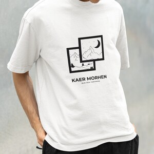 May include: White t-shirt with a black graphic design of a mountain landscape with a crescent moon. The text "Kaer Morhen Old Sea Fortress" is printed below the graphic.