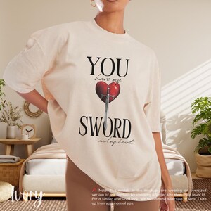 May include: A beige t-shirt with a graphic print of a red heart with a sword through it. The text "YOU have my SWORD and my heart..." is printed on the shirt.