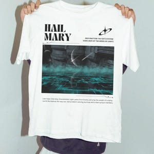 May include: White t-shirt with a graphic design featuring a futuristic cityscape, a blue and green grid, and the text "HAIL MARY" and "DESTINATION TAU CETI SYSTEM 0.920 (92% OF THE SPEED OF LIGHT)".