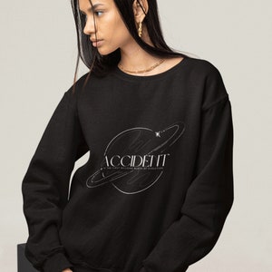 May include: Black sweatshirt with a white graphic design of a planet with rings and the text "ACCIDENT IS THE FIRST BUILDING BLOCK OF EVOLUTION".