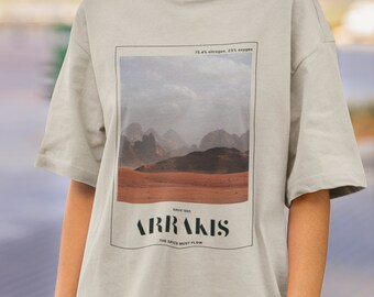 May include: A beige t-shirt with a graphic print of a desert landscape and the text "Arrakis, The Spice Must Flow".