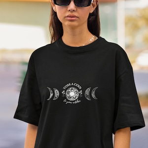 May include: Black t-shirt with a white graphic design featuring three crescent moons and the text "Toss a coin to your witcher".