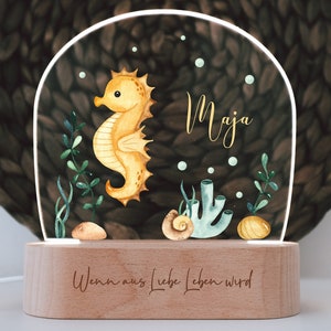 Peut inclure: Veilleuse décorative avec un motif d'hippocampe et le nom "Maja" en lettres dorées. La lumière a une base en bois avec le texte "Wenn aus Liebe Leben wird". L'hippocampe est jaune et le fond a un thème sous-marin.