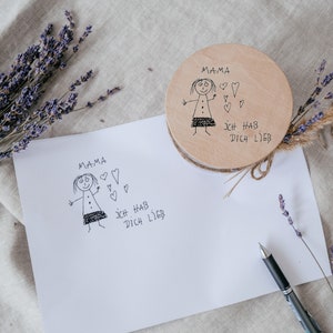 May include: A white card with a hand-drawn illustration of a girl and the text "Mama Ich Hab Dich Lieb" on a light brown linen background. A wooden round box with the same illustration and text is also visible.
