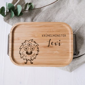 Op de afbeelding: Een houten lunchbox met een schattig monsterontwerp en de tekst "Krümelmonster Levi" erop gegraveerd.