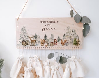 May include: Wooden Advent calendar with a winter forest scene and the text "Adventskalender von Hanna". The calendar has 24 small fabric bags hanging from it.