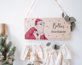 May include: A wooden advent calendar with a white bear dressed as Santa Claus holding a red bag. The calendar has 24 small fabric bags hanging from it. The sign reads "Bilbos Adventskalender".