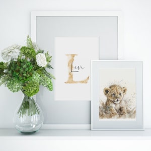 Elegante Bilderrahmen auf einem Regal präsentieren einen zauberhaften Aquarelldruck mit einem niedlichen Löwenbaby. Die sanften Farben und das zarte Design verleihen jedem Raum eine harmonische, naturverbundene Atmosphäre. Daneben steht ein frischer Blumenstrauß in einer Glasvase, der das Arrangement perfekt ergänzt und für einen Hauch von Eleganz sorgt. Ideal für Kinderzimmer oder Wohnzimmer.