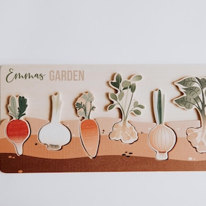 Puede incluir: Rompecabezas de madera con temática de jardín con el texto "Emmas GARDEN". El rompecabezas incluye formas de verduras como rábano, ajo, zanahoria y cebolla. Las verduras son en tonos de rojo, naranja y verde.