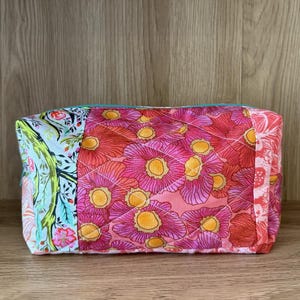 May include: A rectangular quilted cosmetic bag with a floral pattern. The bag features a vibrant mix of colors, including pink, red, and yellow flowers on a light pink background. The bag has a teal zipper and is set against a wooden background.