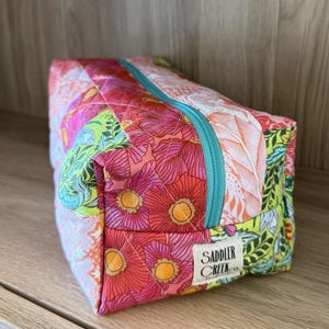 May include: A quilted floral print cosmetic bag with a turquoise zipper. The bag features a patchwork design with pink, red, and green floral patterns. The label reads "Saddler Creek co."