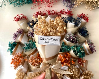 May include: A collection of small bouquets of dried baby's breath flowers wrapped in white paper with gold accents. The bouquets are tied with a gold ribbon and have a small tag with the text "Fabea & Manuel 18.08.2023".